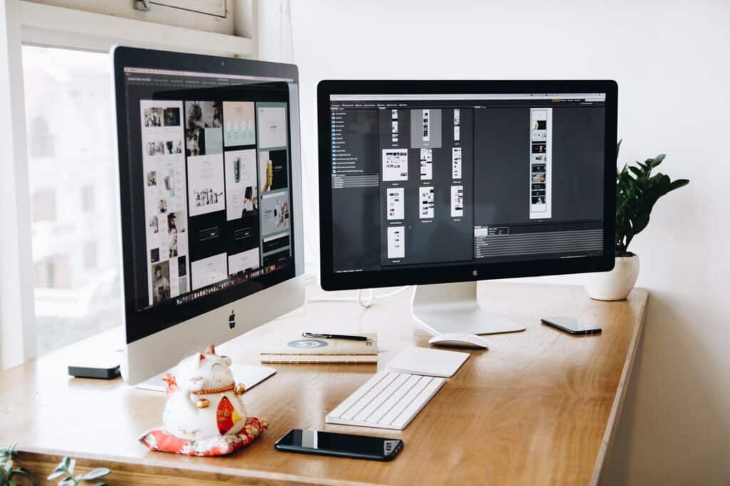 Two Apple iMac computers on a wooden desk with design-related screens displayed. Accessories include a smartphone, notebook, plant, and a Maneki-neko figure.